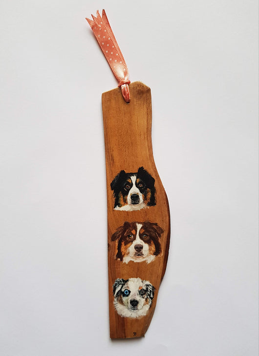 A wooden bookmark with hand-painted portraits of three Australian Shepherd dogs, each with different markings and expressions. The bookmark is tied with a pink ribbon.