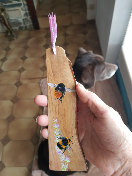 A wooden bookmark with hand-painted illustrations of two bumblebees hovering near flowers. The bookmark is held in a person's hand.