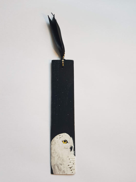 A wooden bookmark with a hand-painted portrait of a snowy owl on a black background. The bookmark is tied with a black ribbon.