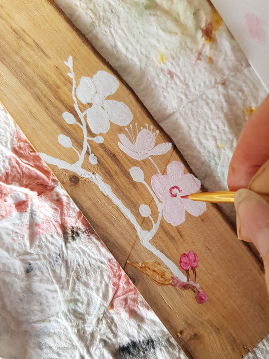 A person's hand painting a cherry blossom on a wooden bookmark with a small paintbrush.